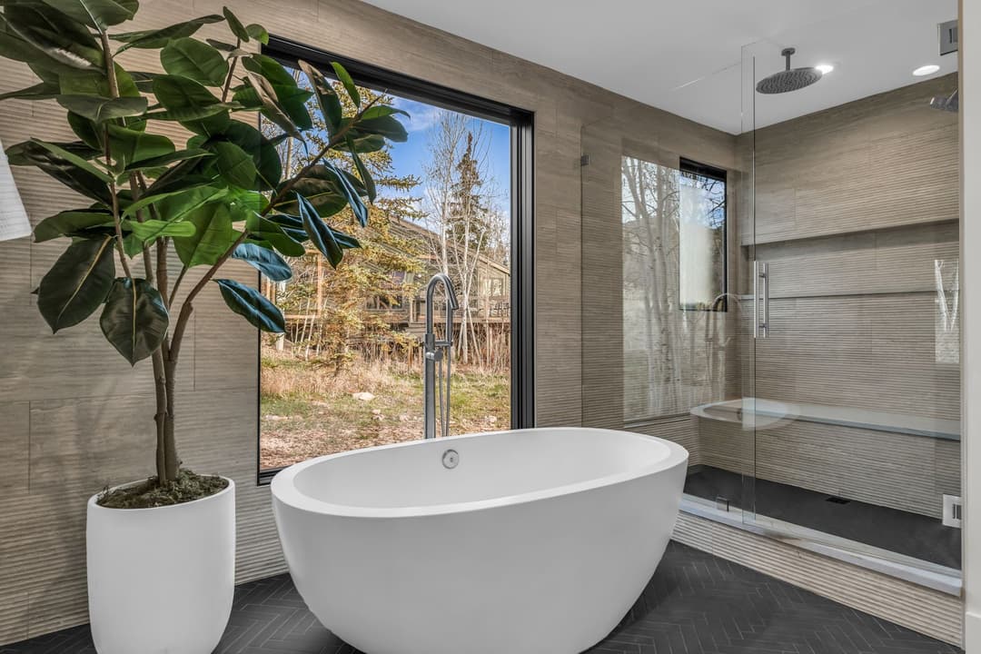 Modern bathroom with freestanding tub, glass shower, and large window overlooking nature.