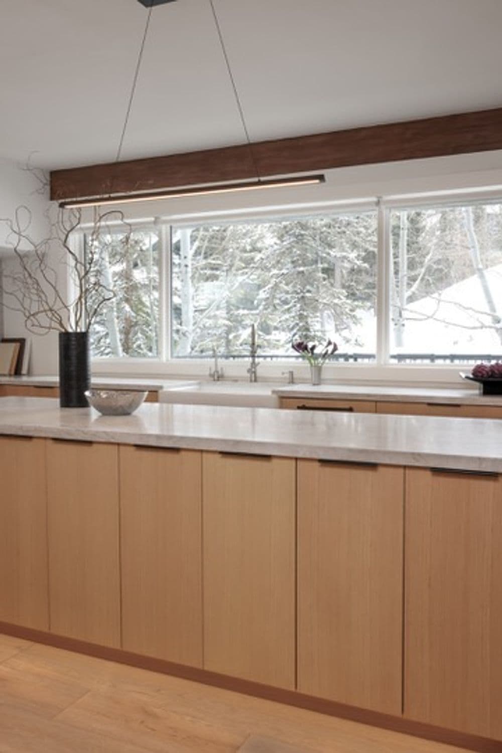 Modern kitchen with wood cabinetry and large windows overlooking a snowy landscape.