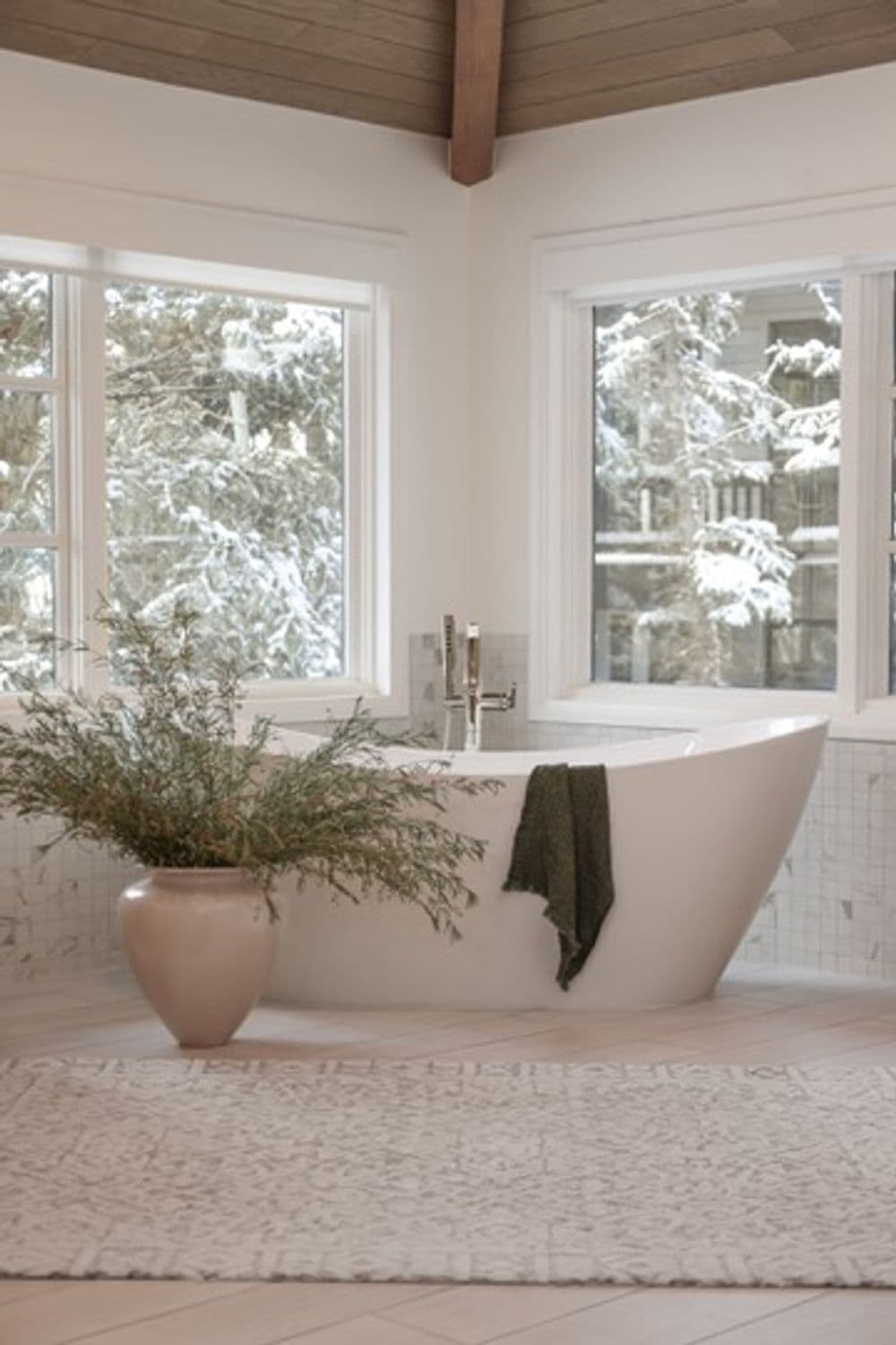 Modern freestanding bathtub with greenery, natural light, and snowy view through large windows.