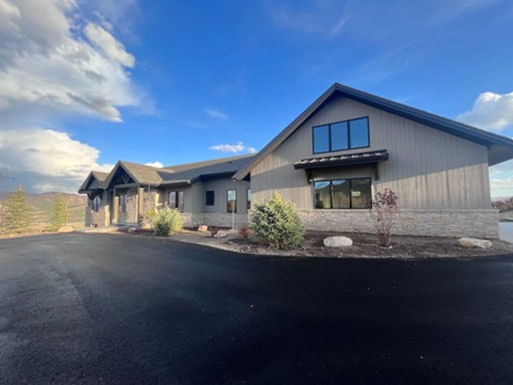 Modern house exterior with stone and wood design, surrounded by landscaped greenery and blue skies.
