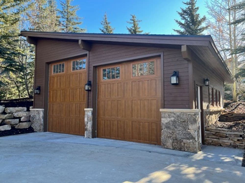 Modern two-car garage with wooden doors, stone accents, and pine trees in the background.