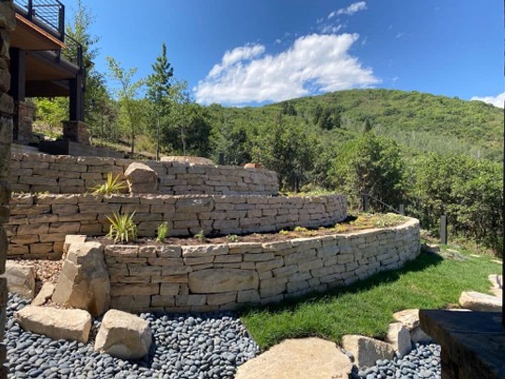 Terraced stone garden with green plants, surrounded by trees and a mountain backdrop.