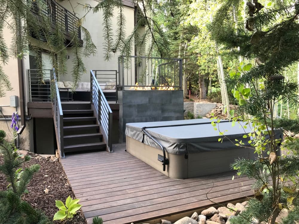 Deck with hot tub and stairs surrounded by greenery and modern home architecture.