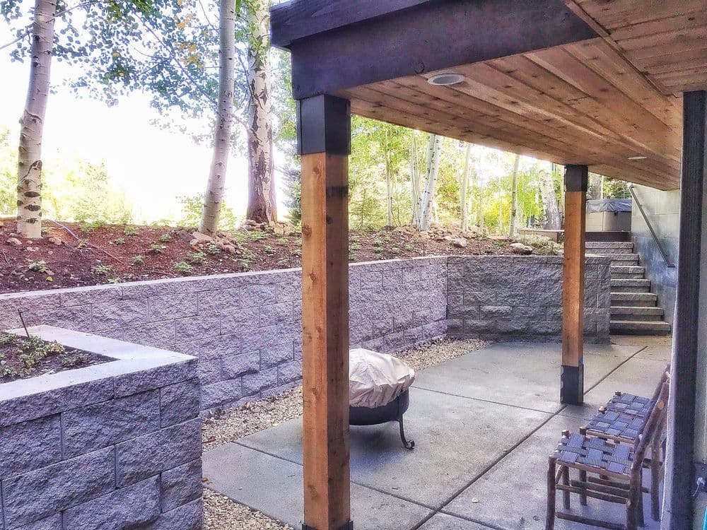 Patio area under wooden structure with stone walls, fire pit, and surrounding trees.
