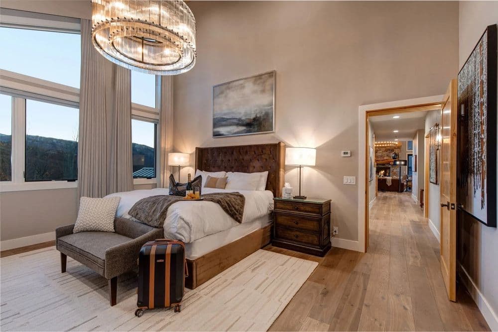 Modern bedroom with large windows, chandelier, and cozy decor, featuring a suitcase and armchair.