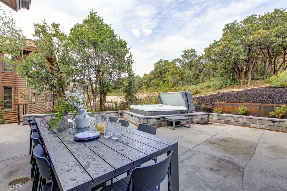 Outdoor patio with a dining table, hot tub, and lush green trees in the background.