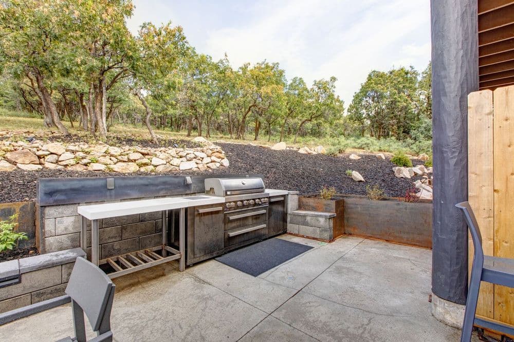 Outdoor kitchen with stainless steel grill and countertop in a lush, wooded setting.